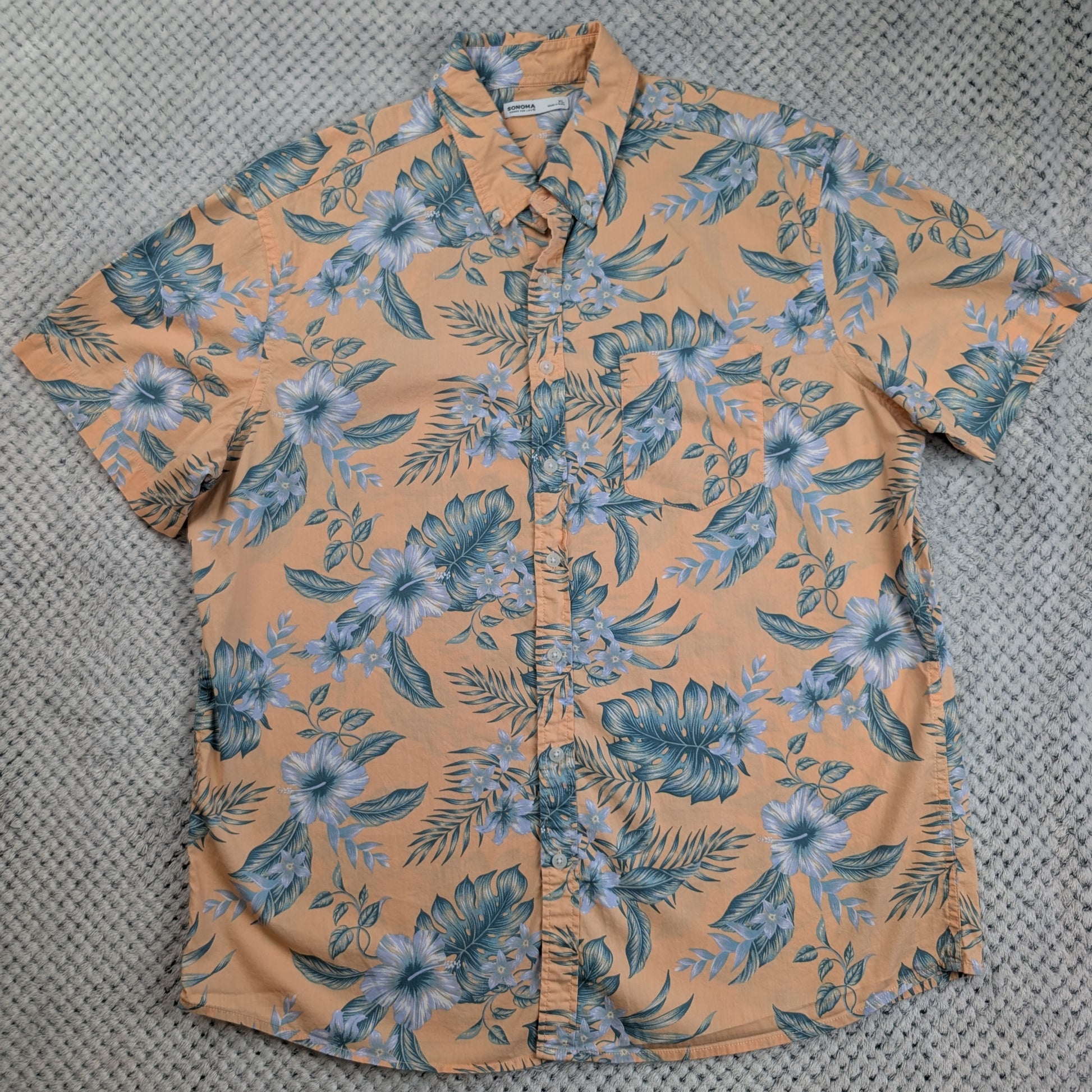 A short-sleeve button-up shirt with a peach-orange base and a blue-green tropical floral print, laid flat on a gray textured surface.