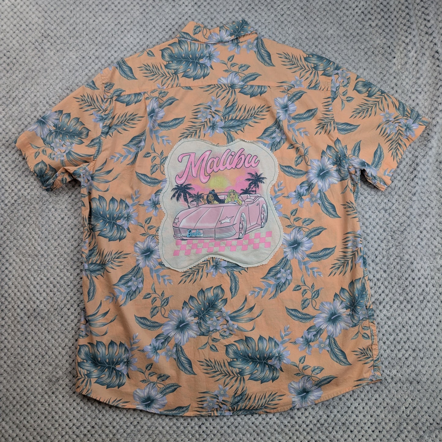 A peach-colored short-sleeve Hawaiian shirt with a blue and white floral print, featuring a large 'Malibu' graphic patch on the back with a pink car and palm trees, laid flat on a gray textured surface.
