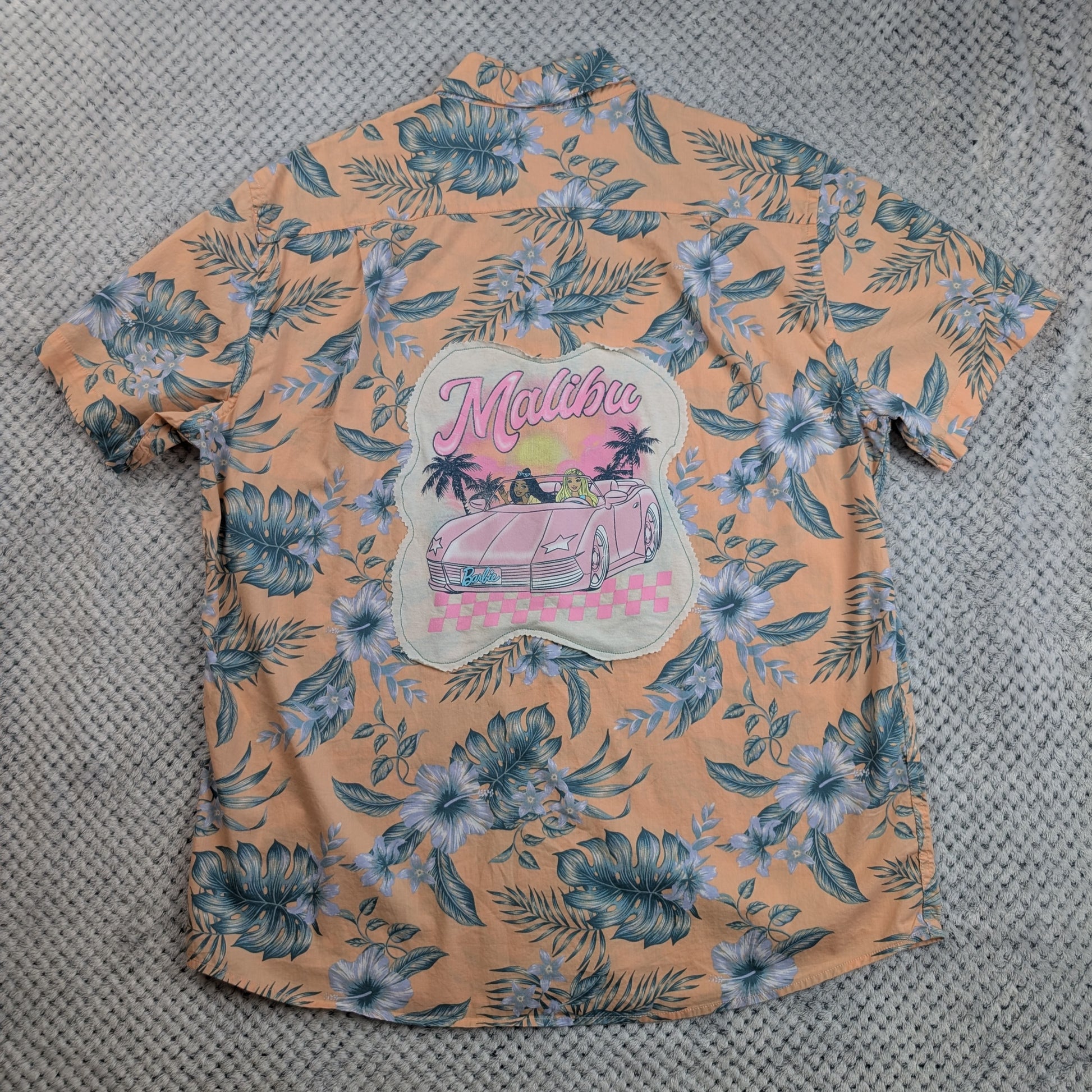A peach-colored short-sleeve Hawaiian shirt with a blue and white floral print, featuring a large 'Malibu' graphic patch on the back with a pink car and palm trees, laid flat on a gray textured surface.