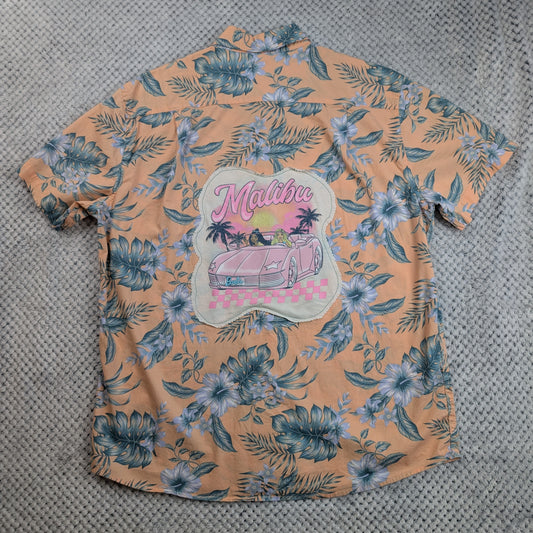 A peach-colored short-sleeve Hawaiian shirt with a blue and white floral print, featuring a large 'Malibu' graphic patch on the back with a pink car and palm trees, laid flat on a gray textured surface.