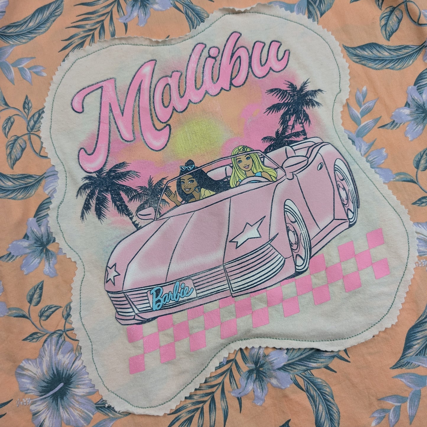 A close-up of a floral fabric featuring a scalloped applique patch with a vintage-style illustration of a pink convertible car, palm trees, and the words 'Malibu' and 'Barbie'.
