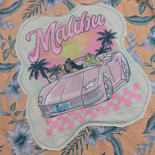 A close-up of a floral fabric featuring a scalloped applique patch with a vintage-style illustration of a pink convertible car, palm trees, and the words 'Malibu' and 'Barbie'.
