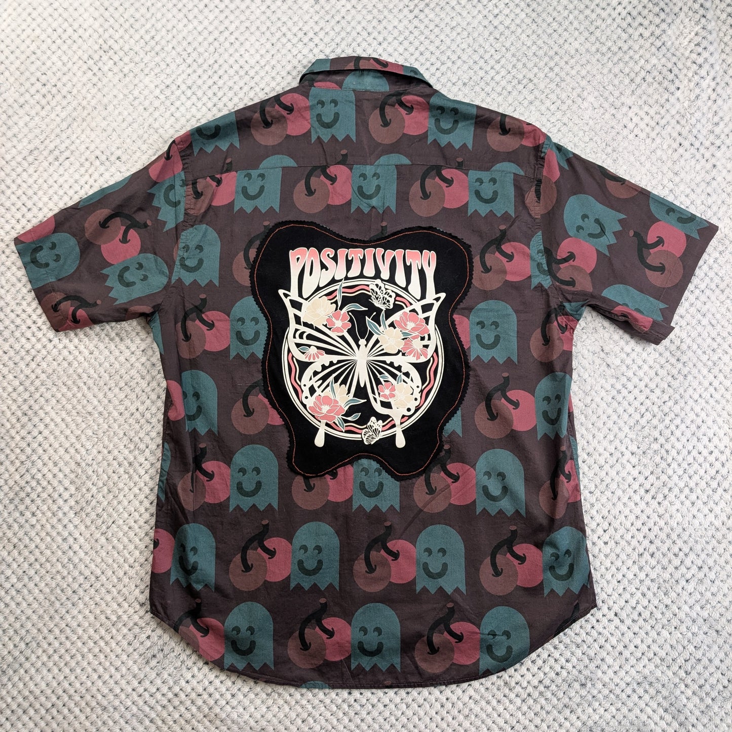 A short-sleeve button-up shirt laid flat on a textured light gray surface, showing the back side. The shirt has an allover print of pink and teal ghosts on a dark brown background, with a large black graphic patch in the center featuring a butterfly, flowers, and the word 'Positivity'.