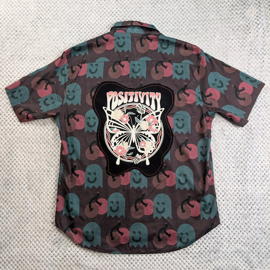 A short-sleeve button-up shirt laid flat on a textured light gray surface, showing the back side. The shirt has an allover print of pink and teal ghosts on a dark brown background, with a large black graphic patch in the center featuring a butterfly, flowers, and the word 'Positivity'.