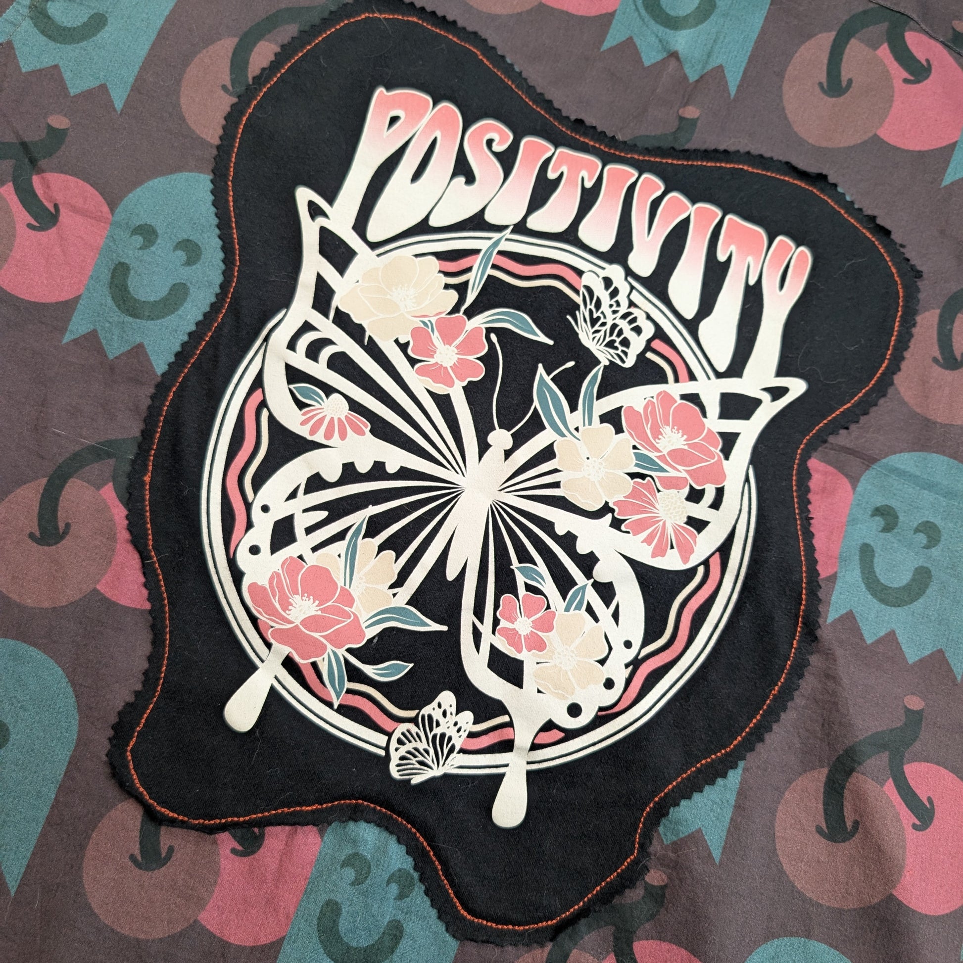 A black fabric patch with a scalloped edge featuring a butterfly and floral design with the word 'POSITIVITY' in pink and white, laid flat on a patterned fabric background.
