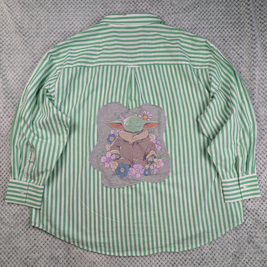 A green and white vertically striped long-sleeve button-up shirt laid flat on a textured gray background, featuring a large graphic print of Grogu/Baby Yoda surrounded by flowers on the back.