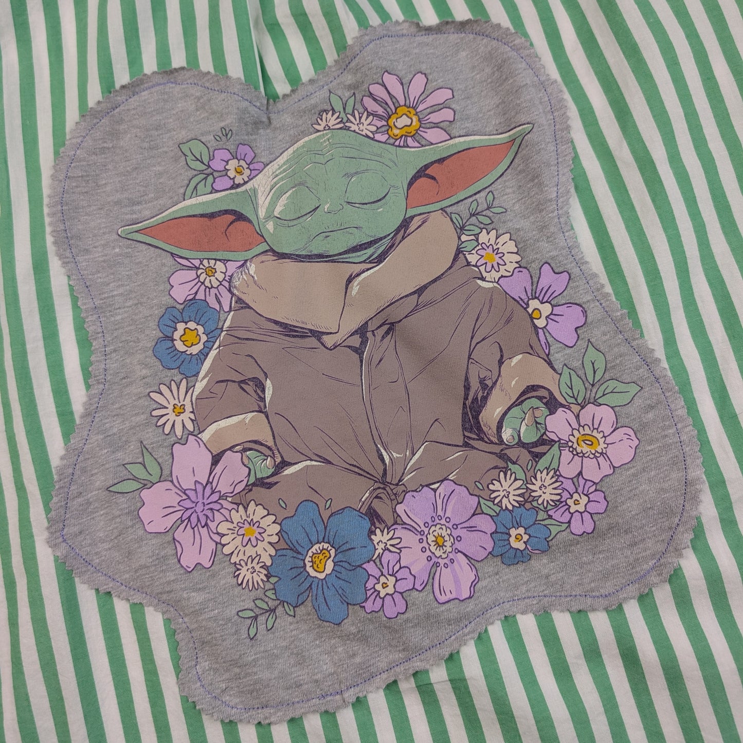 A gray fabric patch featuring a sleeping green cartoon character surrounded by purple, blue, and white flowers, placed on a green and white striped background.