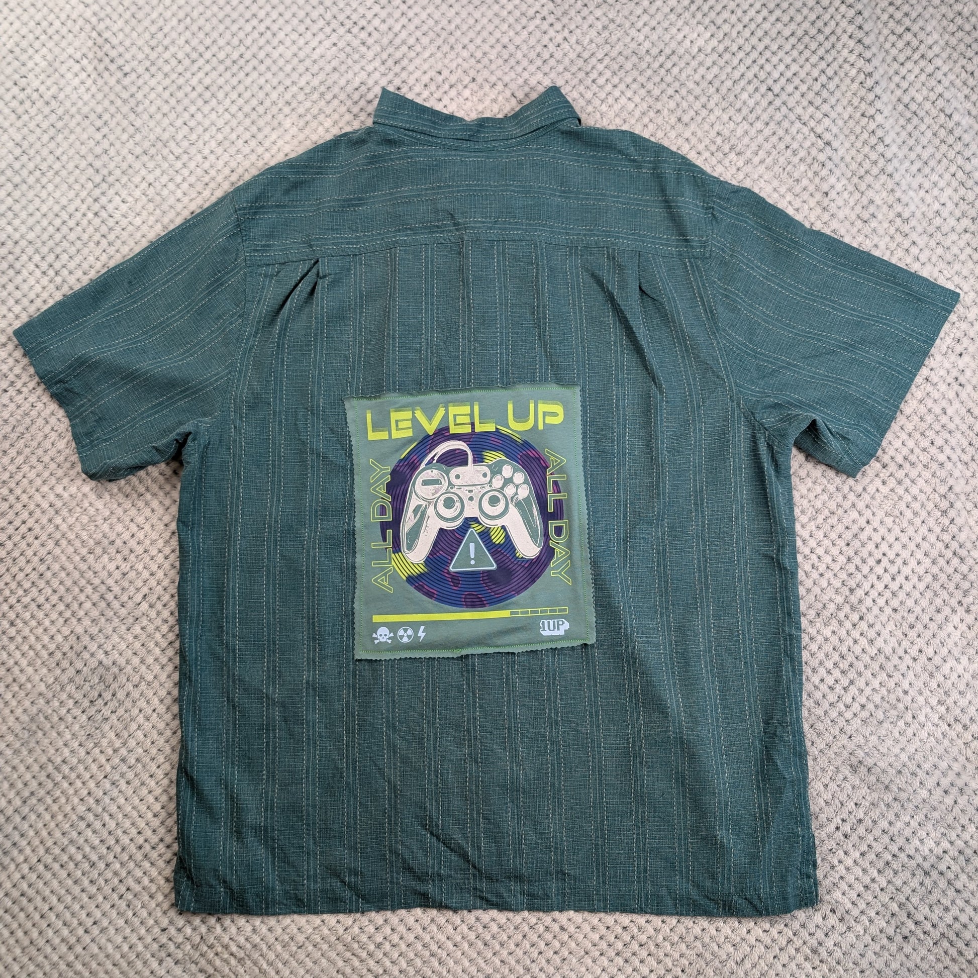 A dark teal green short-sleeve button-up shirt with thin vertical pinstripes, laid flat on a textured beige knit blanket. The back of the shirt features a large graphic patch with the text 'LEVEL UP' in yellow, a white video game controller, and a purple circular background.