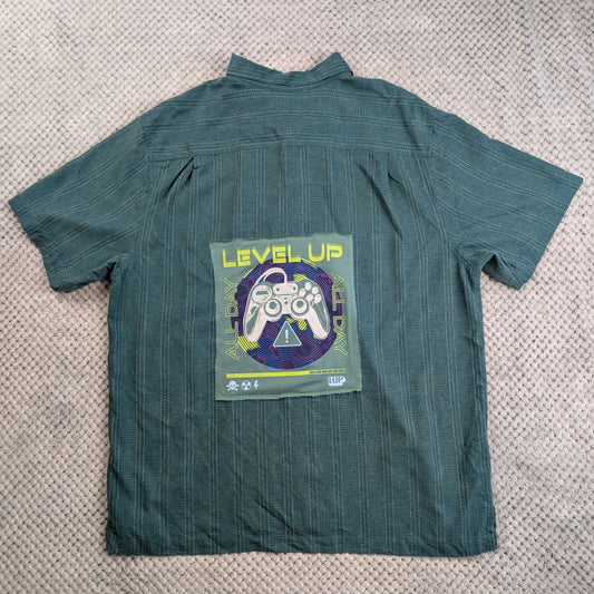 A dark teal green short-sleeve button-up shirt with thin vertical pinstripes, laid flat on a textured beige knit blanket. The back of the shirt features a large graphic patch with the text 'LEVEL UP' in yellow, a white video game controller, and a purple circular background.