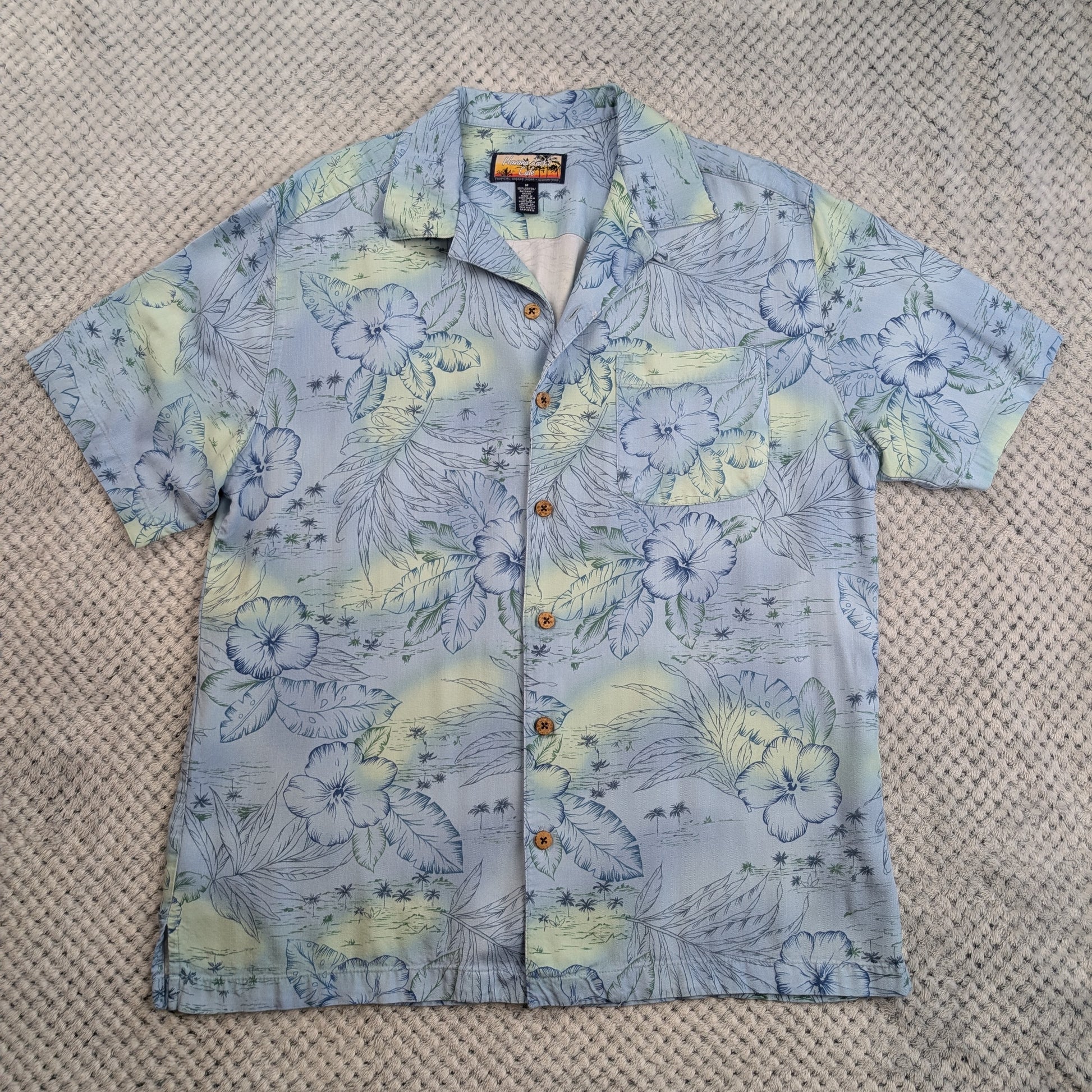 A short-sleeve button-up Hawaiian shirt with a blue and yellow tropical floral print, laid flat on a textured beige woven rug.