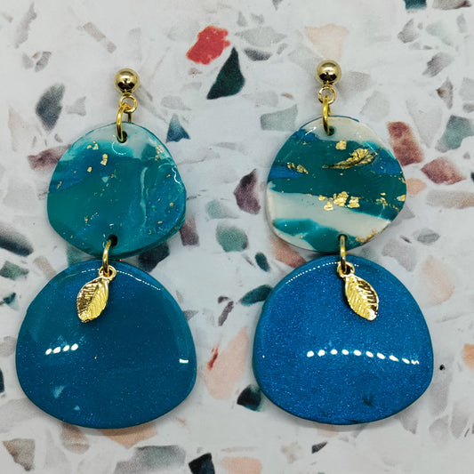 Blue and green earrings with gold leaf details on a patterned surface