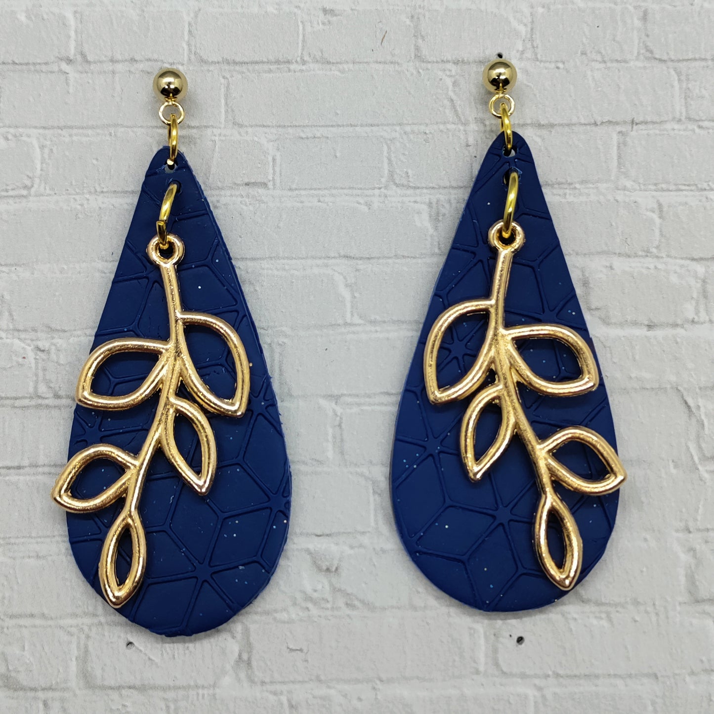 Navy blue earrings with gold leaf designs on a light gray brick wall background