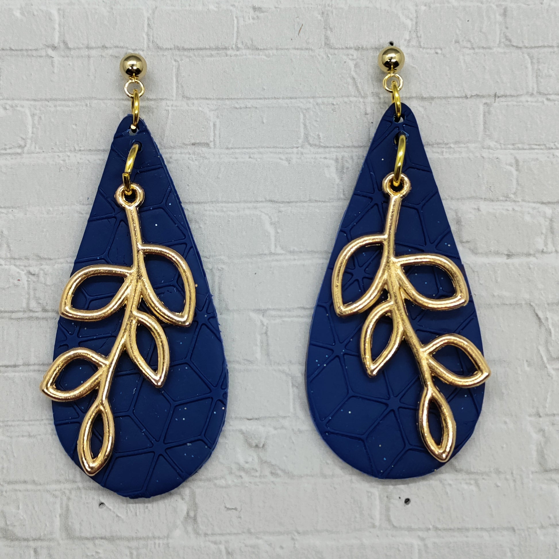 Navy blue earrings with gold leaf designs on a light gray brick wall background