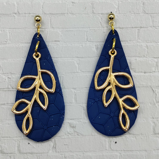 Navy blue earrings with gold leaf designs on a light gray brick wall background
