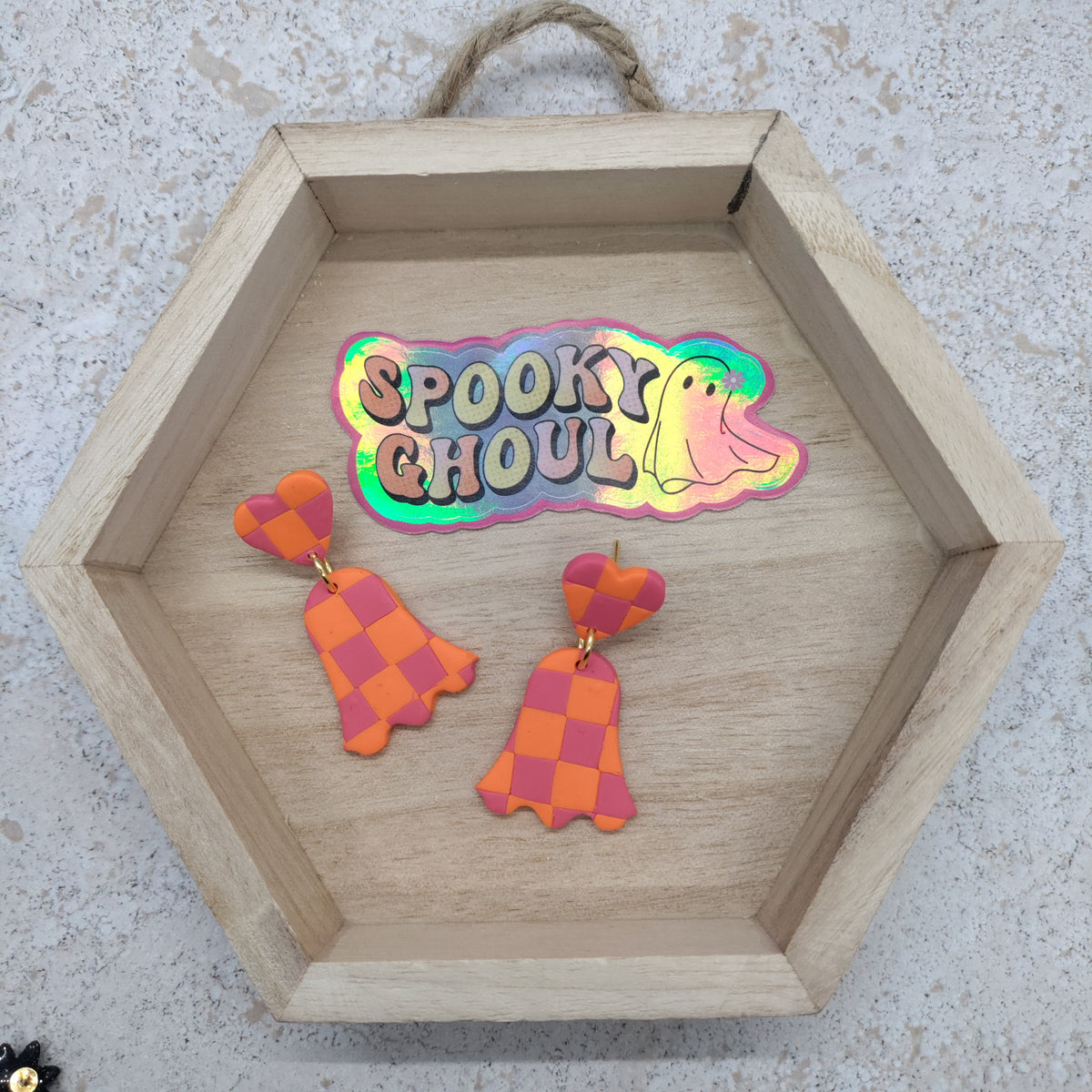 A pair of orange and pink checkered bell-shaped dangle earrings displayed inside a hexagonal wooden tray with a holographic 'Spooky Ghoulish' sticker on a textured light gray background.
