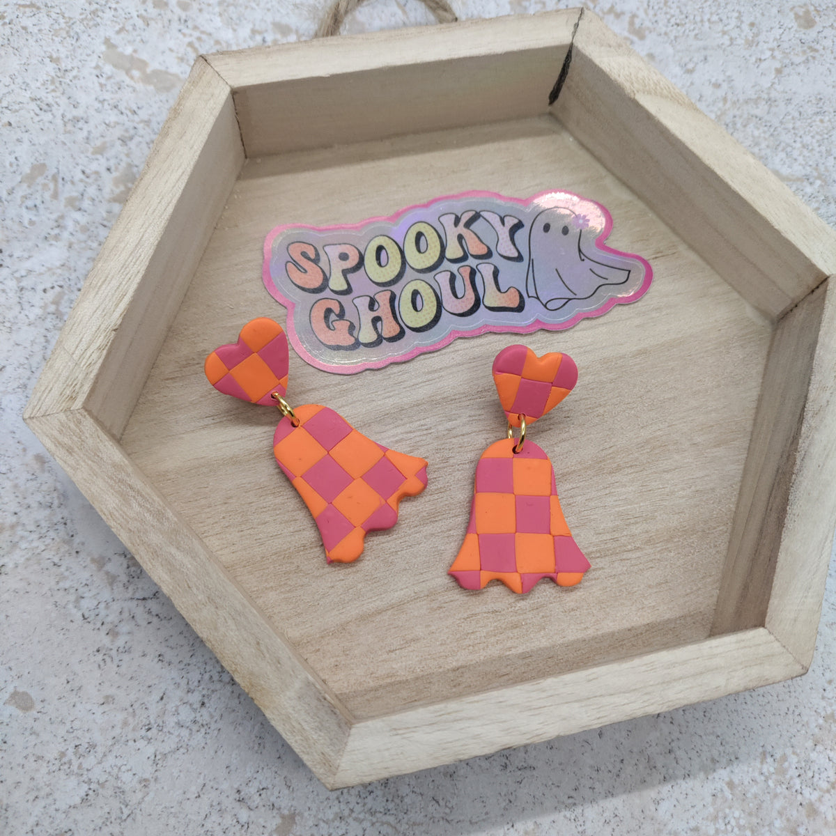 A pair of orange and pink checkered ghost drop earrings displayed on a light wood hexagonal tray next to a 'Spooky Ghoulish' sticker.