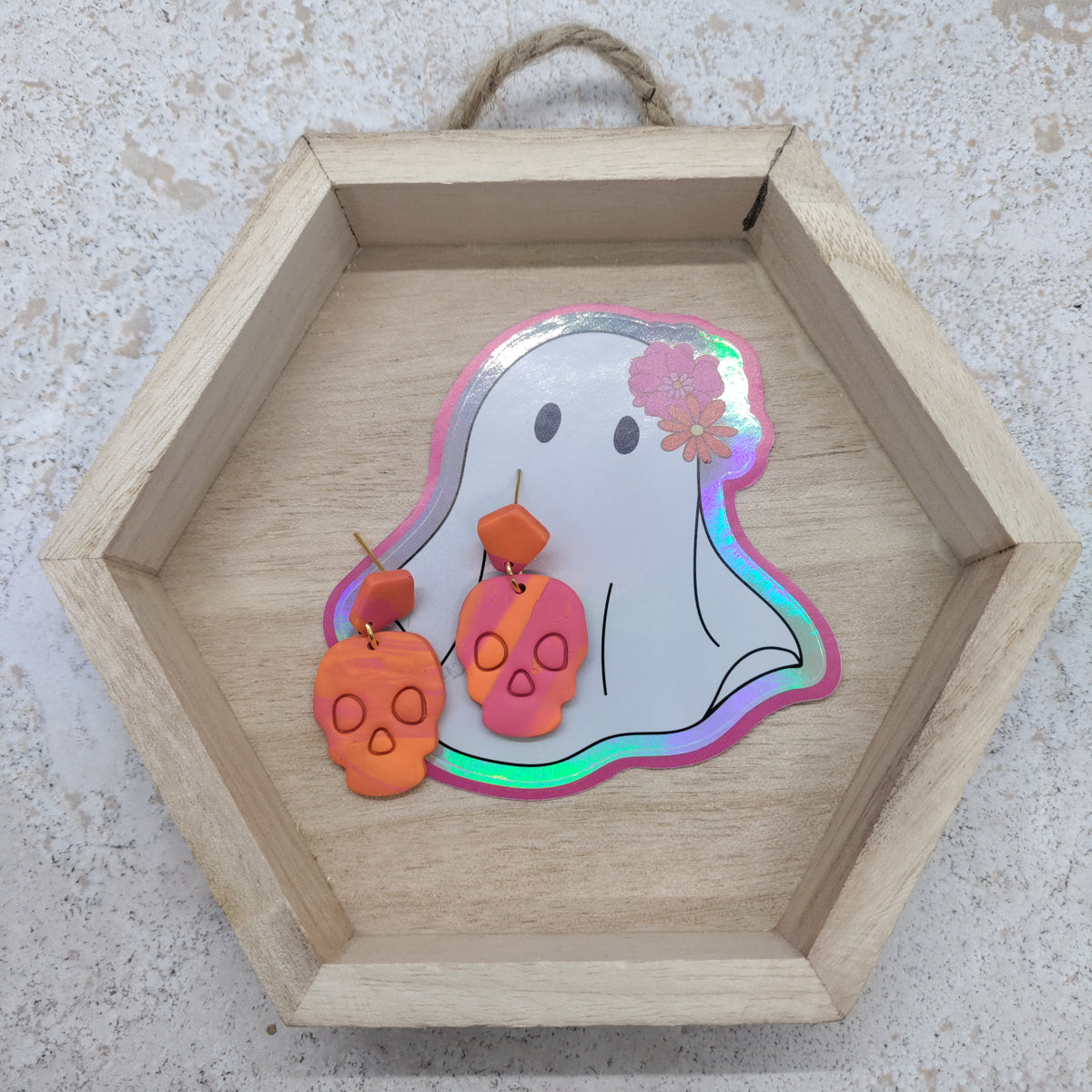 Halloween-themed earrings displayed inside a hexagonal wooden tray. The earrings feature two orange skull charms with dangling orange beads and a pink skull charm with a matching pink bead, both adorned with small silver paper wings.