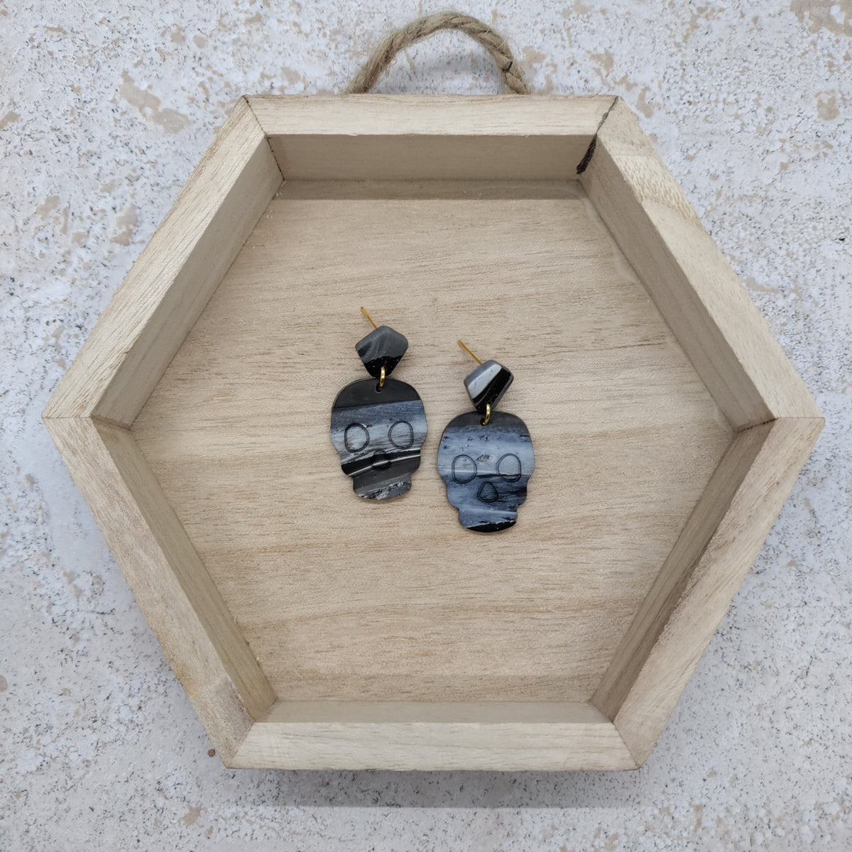 A pair of handmade dangle earrings made from polished black and gray agate slices with gold posts, displayed inside a light wood hexagonal tray on a textured light gray surface.