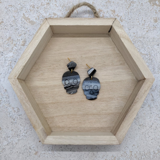 A pair of handmade dangle earrings made from polished black and gray agate slices with gold posts, displayed inside a light wood hexagonal tray on a textured light gray surface.