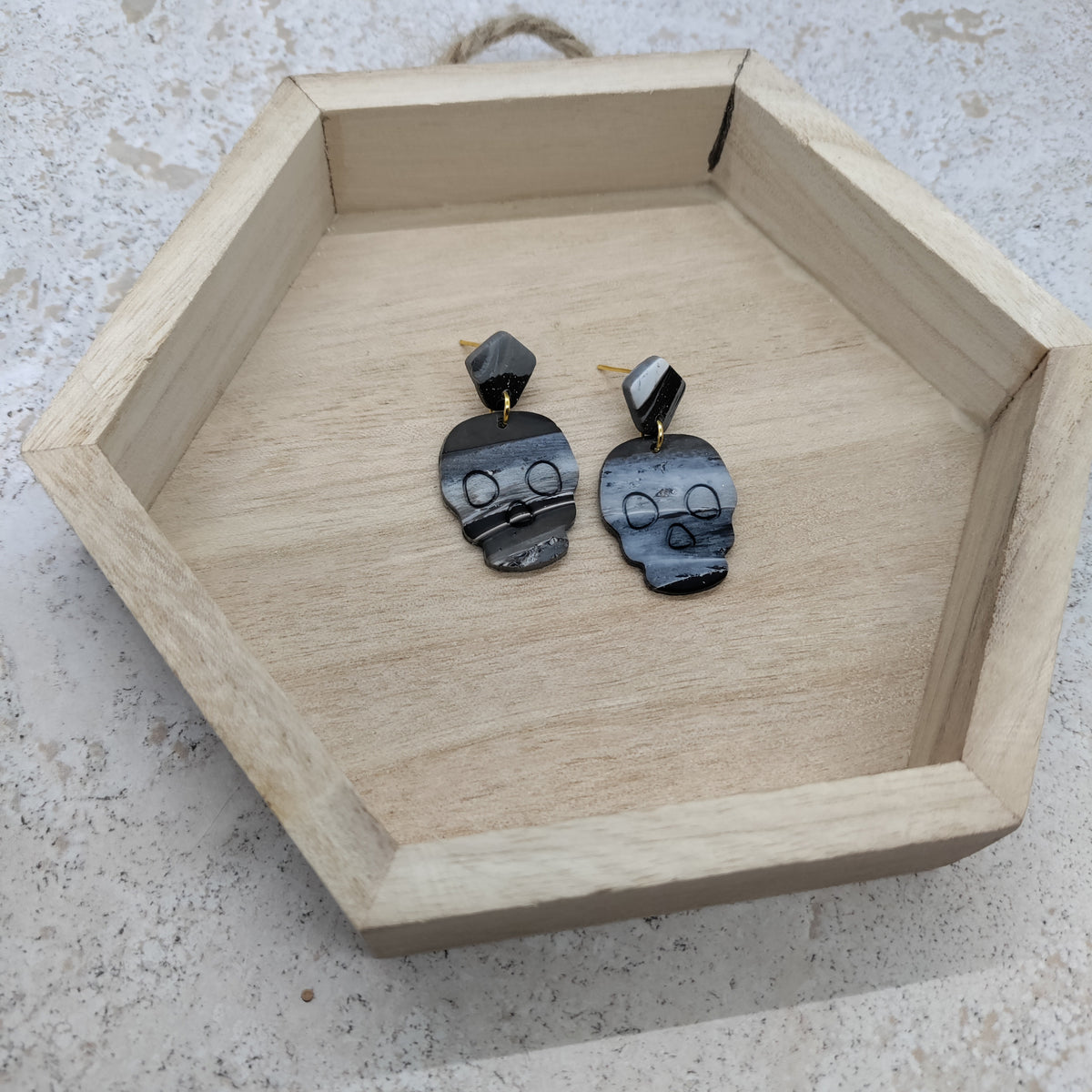 A pair of black skull-shaped drop earrings with gold-toned posts, displayed inside a light wood hexagonal tray on a speckled stone surface.