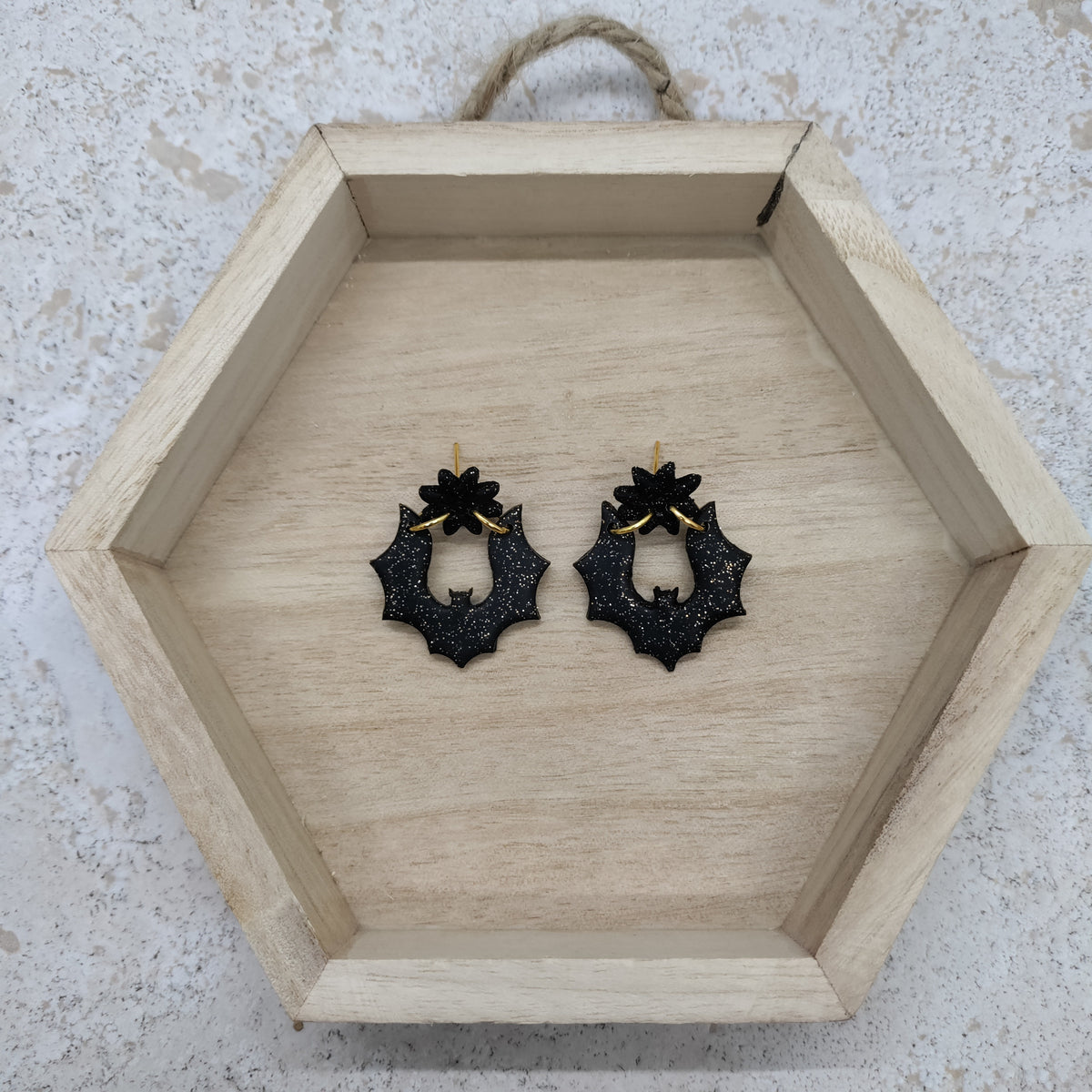 A pair of black holly leaf dangle earrings with gold posts, displayed inside a hexagonal wooden tray on a textured light gray surface.