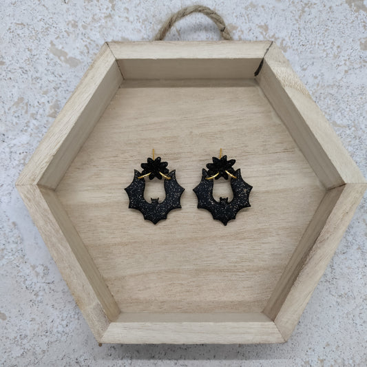 A pair of black holly leaf dangle earrings with gold posts, displayed inside a hexagonal wooden tray on a textured light gray surface.