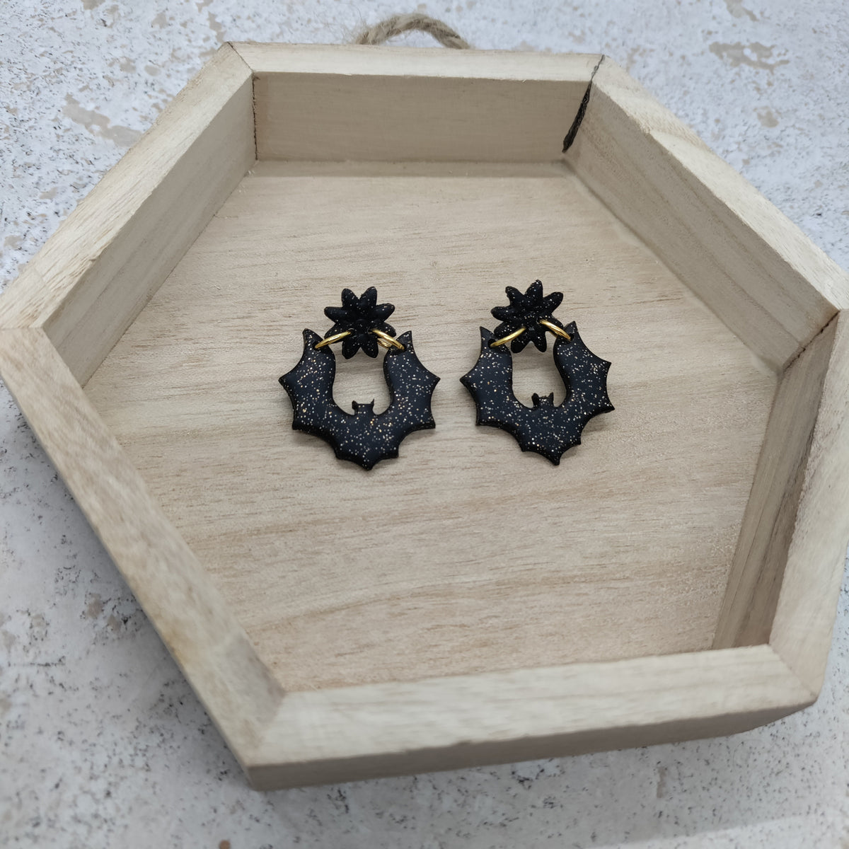 A pair of black bat-shaped hoop earrings with gold-tone wire hangers, displayed inside a hexagonal wooden tray on a light stone surface.