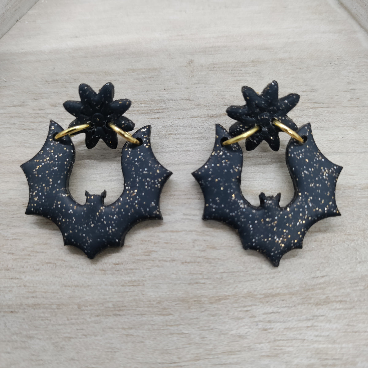 A pair of black bat-shaped hoop earrings with gold-tone ear loops and a small gold star detail on top, displayed on a light wood background.