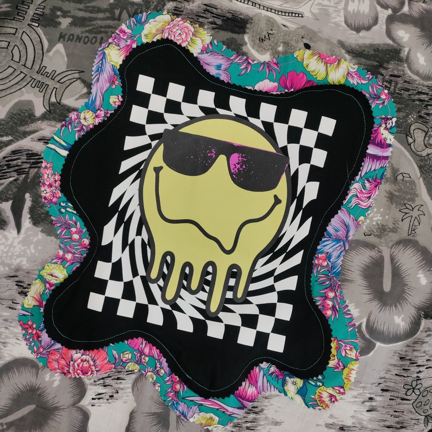 Decorative patch with a smiley face wearing sunglasses on a checkered background, surrounded by colorful floral patterns on a textured surface.