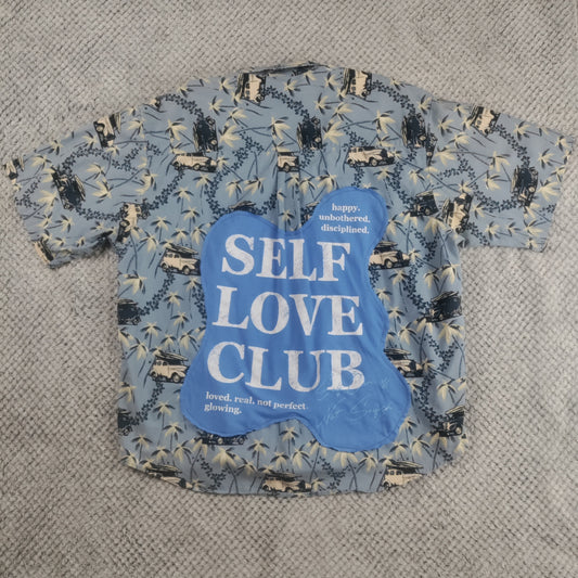 A short-sleeve t-shirt with a blue, beige, and black abstract pattern, featuring a large blue graphic on the front that reads 'SELF LOVE CLUB' in white text, laid flat on a gray textured blanket.