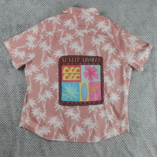 A pink short-sleeve button-up shirt with a white palm tree pattern, featuring a colorful 'SUNLIT SHORES' patch on the back, laid flat on a gray textured blanket.