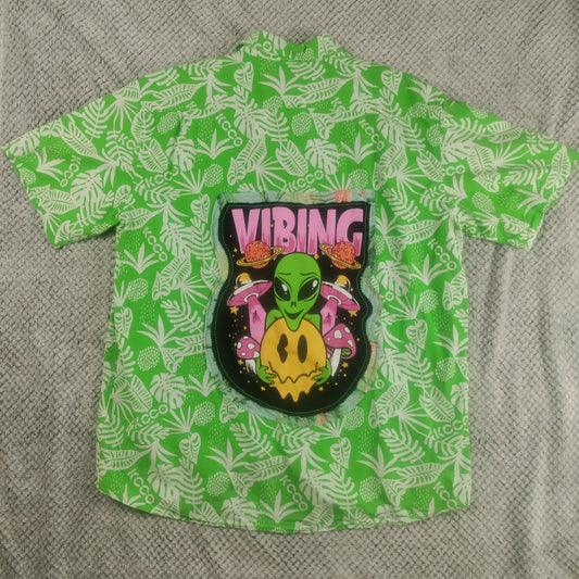 A green short-sleeve button-up shirt with a white tropical leaf pattern, featuring a large central graphic of a green alien with pink sunglasses and the word 'VIBING' in pink above it, laid flat on a gray textured blanket.