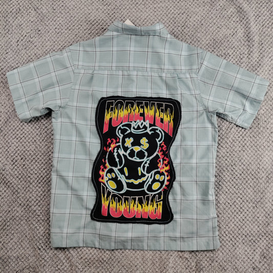 A light gray and white plaid short-sleeve button-up shirt laid flat on a textured gray blanket, featuring a large 'Forever Young' teddy bear patch on the back.