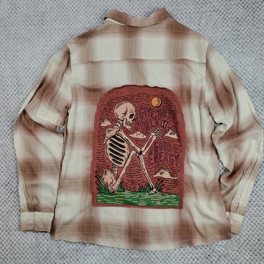 A long-sleeve plaid button-up shirt laid flat on a textured gray surface, showing the back side with a large graphic print of a skeleton over a tombstone and the words 'Give The World Good Energy'.