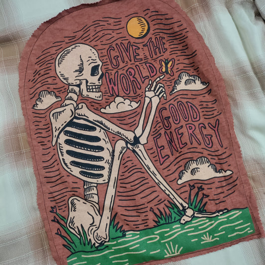 A rectangular fabric patch with a rustic, hand-drawn design of a skeleton sitting on grass under a yellow sun, with the text 'GIVE THE WORLD GOOD ENERGY' on a dusty rose background, laid flat on a beige plaid fabric.