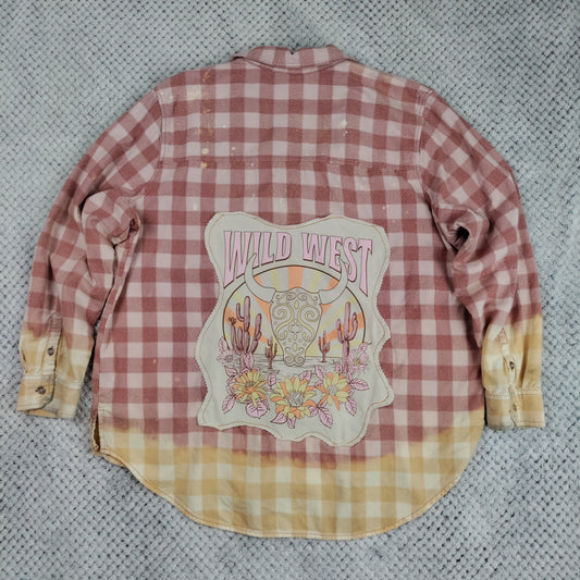 A long-sleeve flannel shirt laid flat on a gray textured blanket. The shirt features a pink and white gingham pattern on the upper body, bleached into a pale yellow gingham at the hem and cuffs.