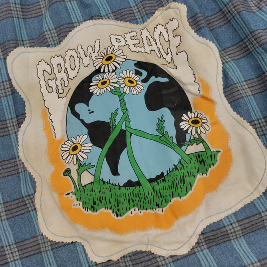 A square fabric patch with a scalloped cream border featuring a peace symbol made from daisy stems over a blue and black globe, with green grass and the words 'GROW PEACE' in white, placed on a blue plaid fabric background.