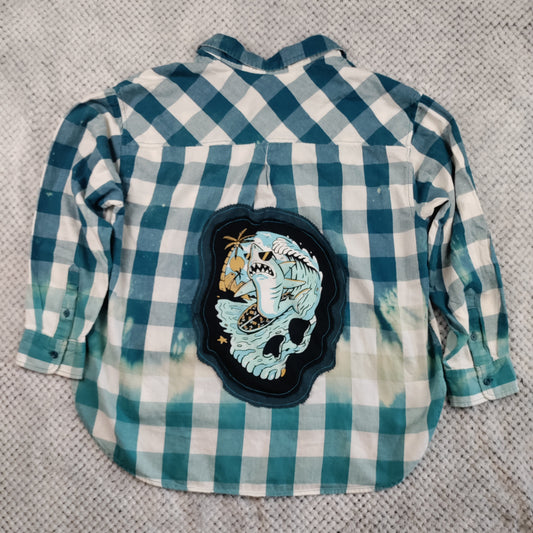 A long-sleeve gingham button-up shirt in teal and white, laid flat on a textured gray surface. The back of the shirt features a large, decorative embroidered patch depicting a light blue skull with a black outline.