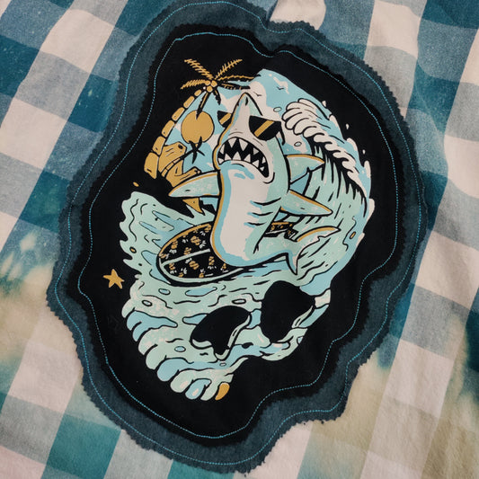 A close-up of a blue and white gingham shirt featuring a large embroidered patch with a shark, skull, and palm tree design.
