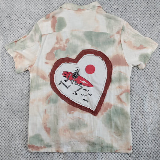 A short-sleeve button-up shirt laid flat on a gray textured blanket. The shirt features a tie-dye pattern in soft beige, sage green, and dusty pink, with a large heart-shaped graphic on the back.