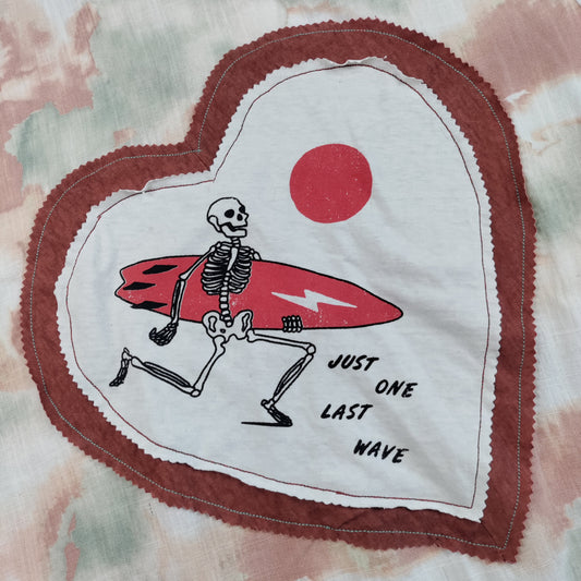 A close-up of a heart-shaped fabric patch sewn onto a tie-dye garment. The patch features a skeleton surfing on a red surfboard with a red sun and the text 'JUST ONE LAST WAVE'.
