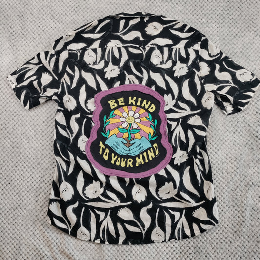 A short-sleeve button-up shirt with a black and white leaf pattern, featuring a colorful graphic patch on the front that reads 'BE KIND TO YOUR MIND' with a flower and hands design, laid flat on a textured gray blanket.