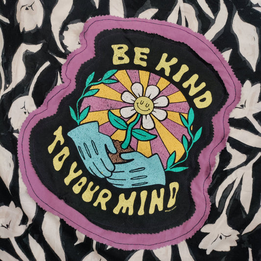 A close-up of a colorful embroidered patch with the text 'BE KIND TO YOUR MIND' surrounding a flower and hands motif, placed on a black and white floral fabric background.