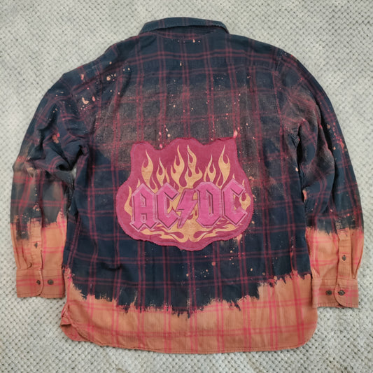 A long-sleeve AC/DC band flannel shirt laid flat on a textured gray fabric background. The back of the shirt features a dark navy and red plaid pattern with a central pink and orange flame-shaped AC/DC logo patch, and a distressed orange-brown tie-dye effect along the bottom hem and cuffs.