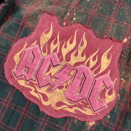 A close-up of a distressed AC/DC band patch with a pink and gold flame design, placed on top of a dark green and red plaid shirt.