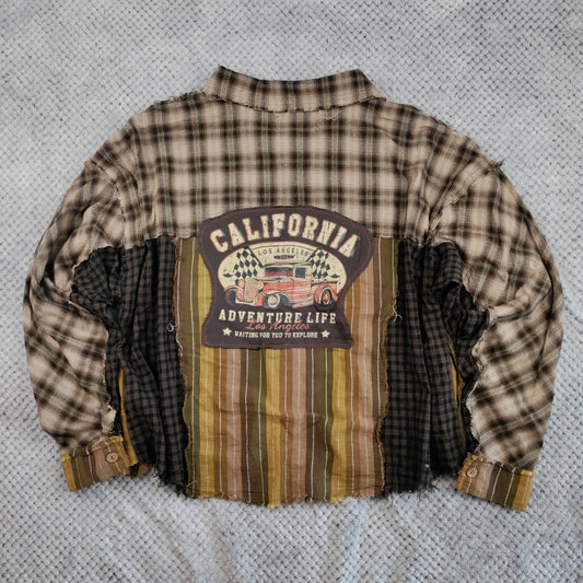 A long-sleeve patchwork flannel shirt laid flat on a textured gray blanket. The shirt features a brown and beige plaid body, a large central patchwork panel with vertical stripes in brown, tan, and olive green, and a dark brown 'CALIFORNIA ADVENTURE LIFE' graphic patch on the chest.