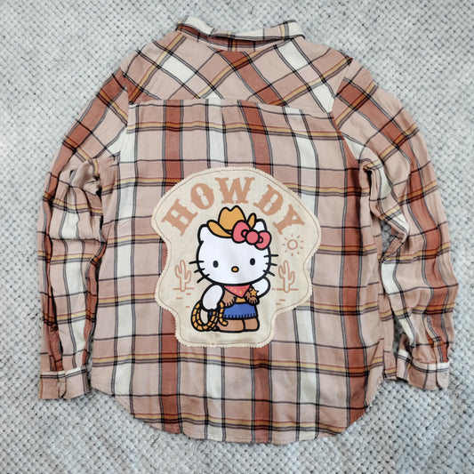 A long-sleeve plaid shirt in beige, brown, and white, featuring a large Hello Kitty cowboy patch on the back with the text 'HOWDY'.