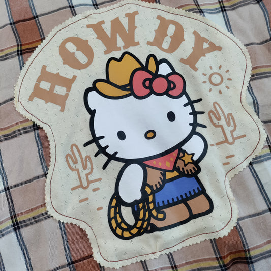 A western-themed Hello Kitty patch with the text 'HOWDY' and a cowboy illustration, placed on a plaid blanket.