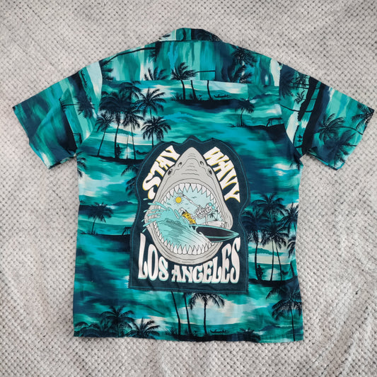 A short-sleeve button-up shirt laid flat on a textured gray blanket. The shirt features a teal and turquoise tropical print with palm trees and a large central graphic of a shark mouth with the words 'Stay Wavy Los Angeles'.