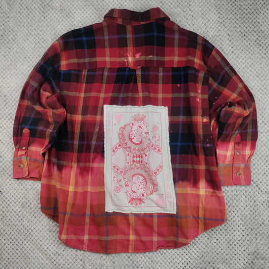 A red and black plaid long-sleeve shirt with a white rectangular patch featuring pink queen of hearts card designs, laid flat on a gray textured blanket.
